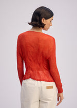 A person with short dark hair is seen from the back, wearing the ISSEY MIYAKE CHIFFON TWIST 2 red textured long-sleeve top and off-white pants with a brown patch, standing against a light gray background.