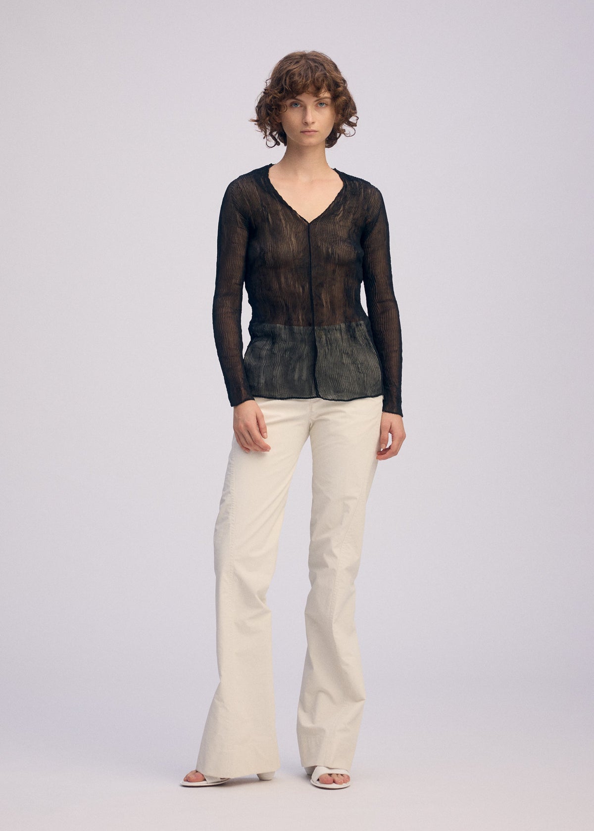 A person with short, curly hair and a neutral expression models the ISSEY MIYAKE CHIFFON TWIST BK/WT-66 sheer V-neck top against a plain background, paired with cream wide-leg pants and white open-toe shoes. view 4 of 6