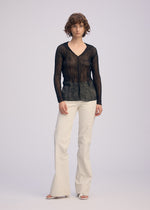 A person with short, curly hair and a neutral expression models the ISSEY MIYAKE CHIFFON TWIST BK/WT-66 sheer V-neck top against a plain background, paired with cream wide-leg pants and white open-toe shoes.