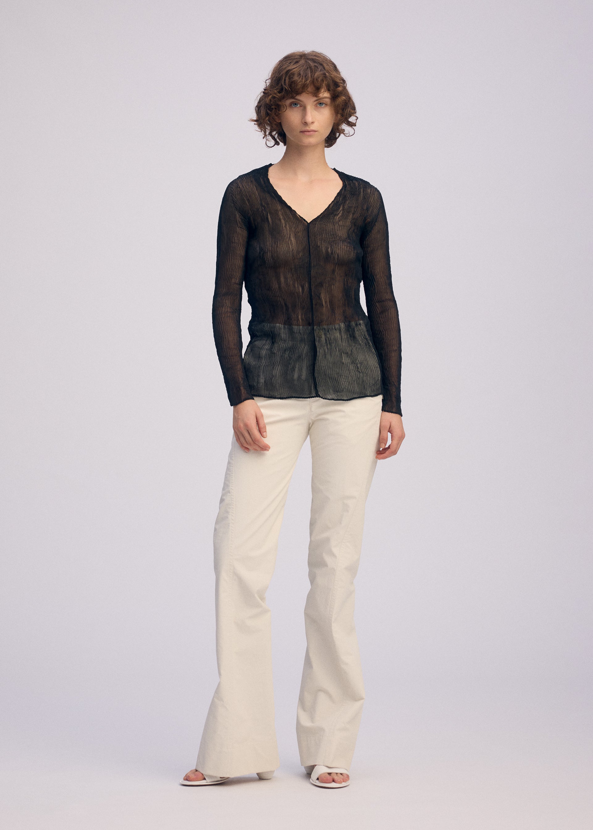 Zoomed view A person with short, curly hair and a neutral expression models the ISSEY MIYAKE CHIFFON TWIST BK/WT-66 sheer V-neck top against a plain background, paired with cream wide-leg pants and white open-toe shoes.