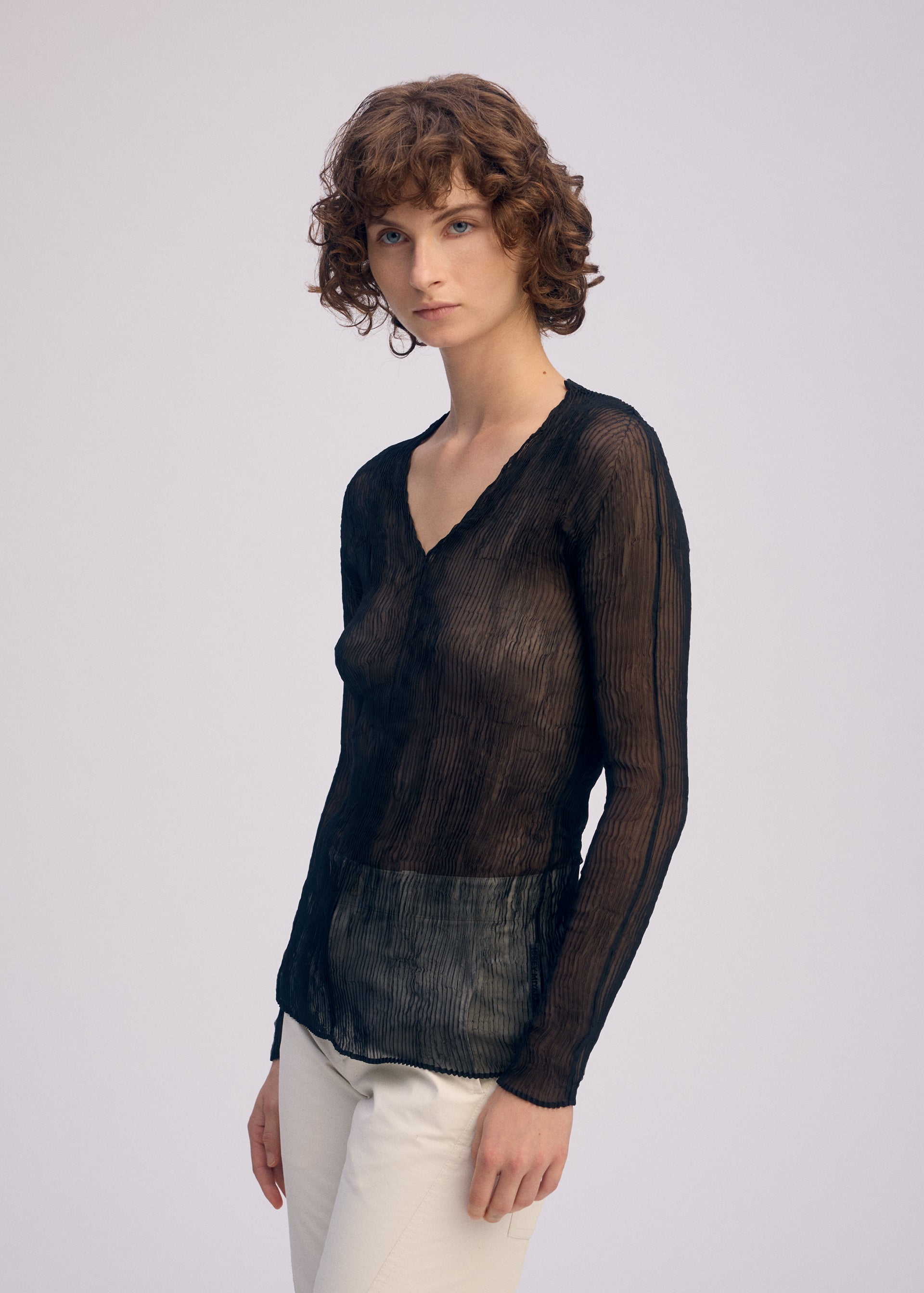 Zoomed view A person with short, curly brown hair wears the ISSEY MIYAKE CHIFFON TWIST BK/WT-66 pleated long-sleeved top and light-colored pants, standing against a plain light background, facing forward with a neutral expression.