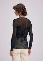 A person with curly hair, seen from the back, wears the CHIFFON TWIST BK/WT-66 sheer V-neck top and light pants by ISSEY MIYAKE, standing against a plain light background. The eco-conscious top is made from recycled polyester.