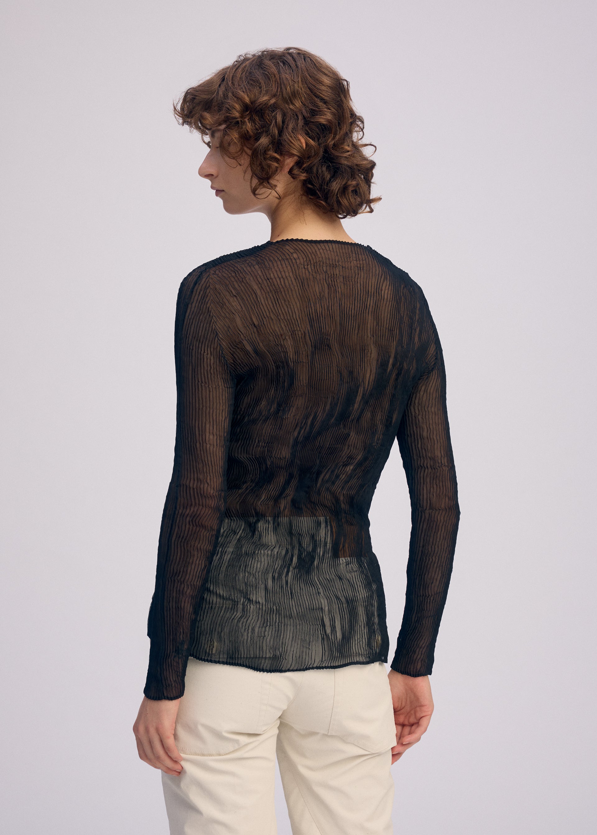 Zoomed view A person with curly hair, seen from the back, wears the CHIFFON TWIST BK/WT-66 sheer V-neck top and light pants by ISSEY MIYAKE, standing against a plain light background. The eco-conscious top is made from recycled polyester.
