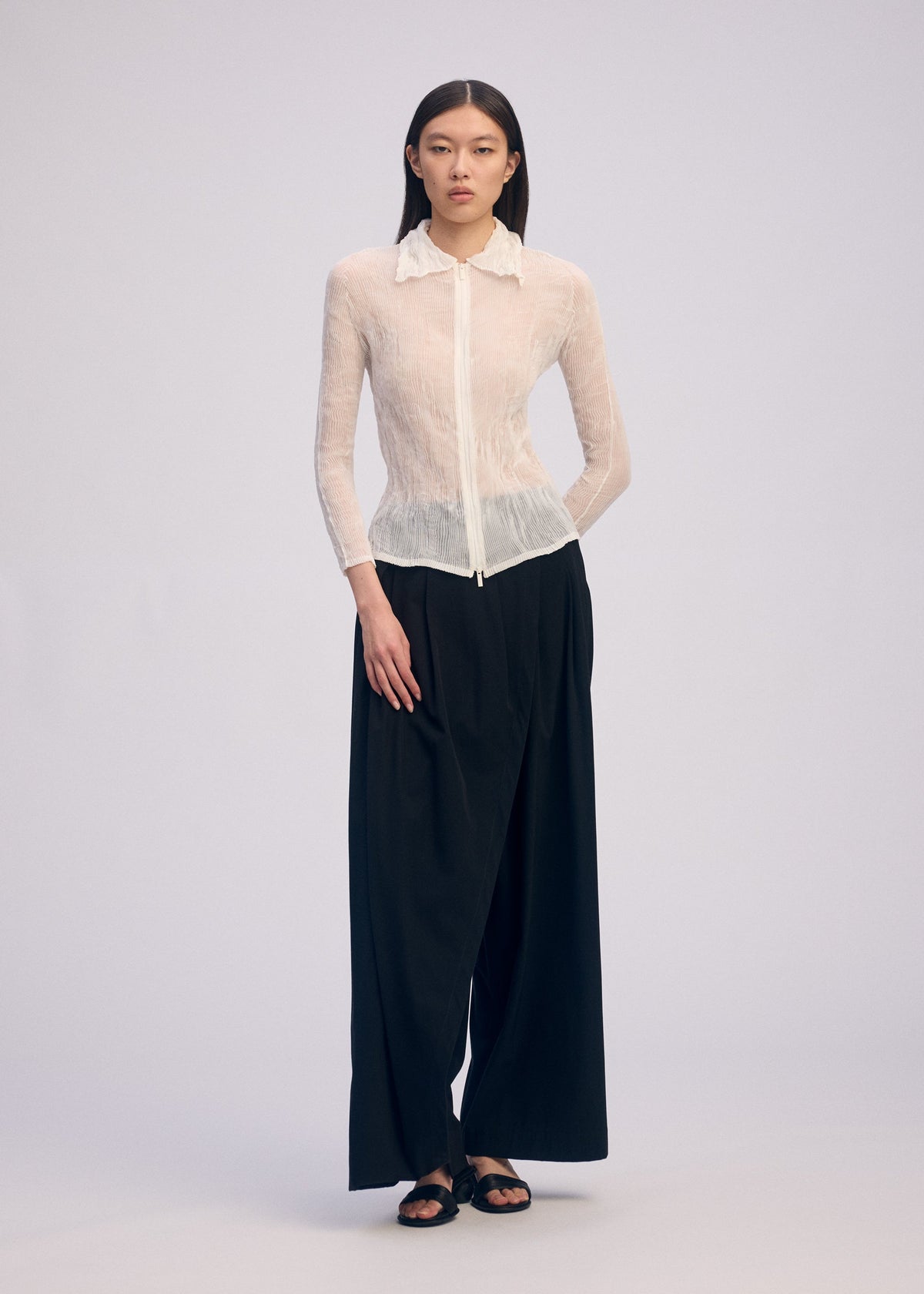 Against a plain light background, a person wears the ISSEY MIYAKE CHIFFON TWIST BK/WT-66: a sheer white long-sleeve collared blouse and pleated black wide-leg pants, paired with black sandals. Their straight hair is worn down. view 4 of 6