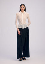 Against a plain light background, a person wears the ISSEY MIYAKE CHIFFON TWIST BK/WT-66: a sheer white long-sleeve collared blouse and pleated black wide-leg pants, paired with black sandals. Their straight hair is worn down.