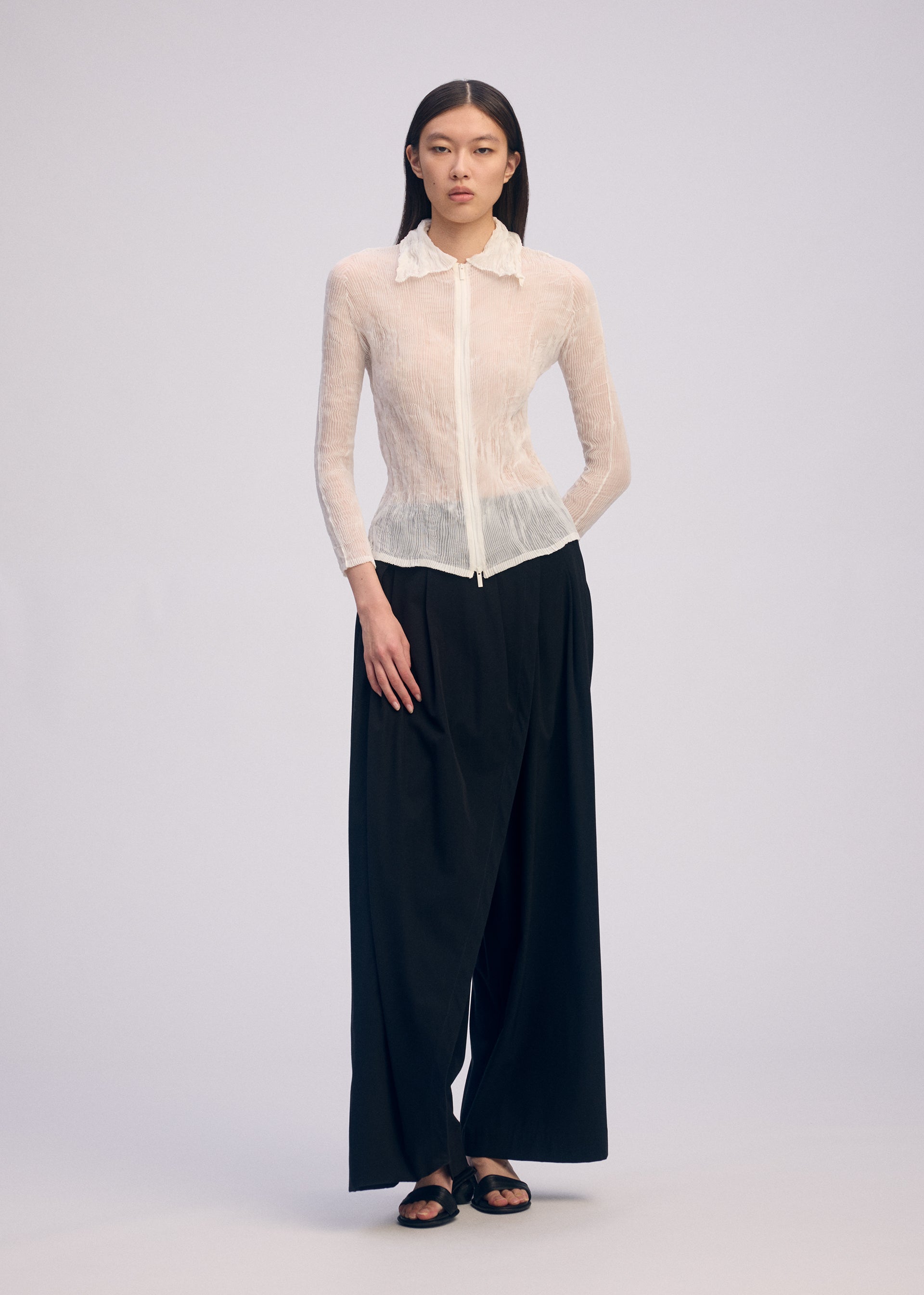 Zoomed view Against a plain light background, a person wears the ISSEY MIYAKE CHIFFON TWIST BK/WT-66: a sheer white long-sleeve collared blouse and pleated black wide-leg pants, paired with black sandals. Their straight hair is worn down.