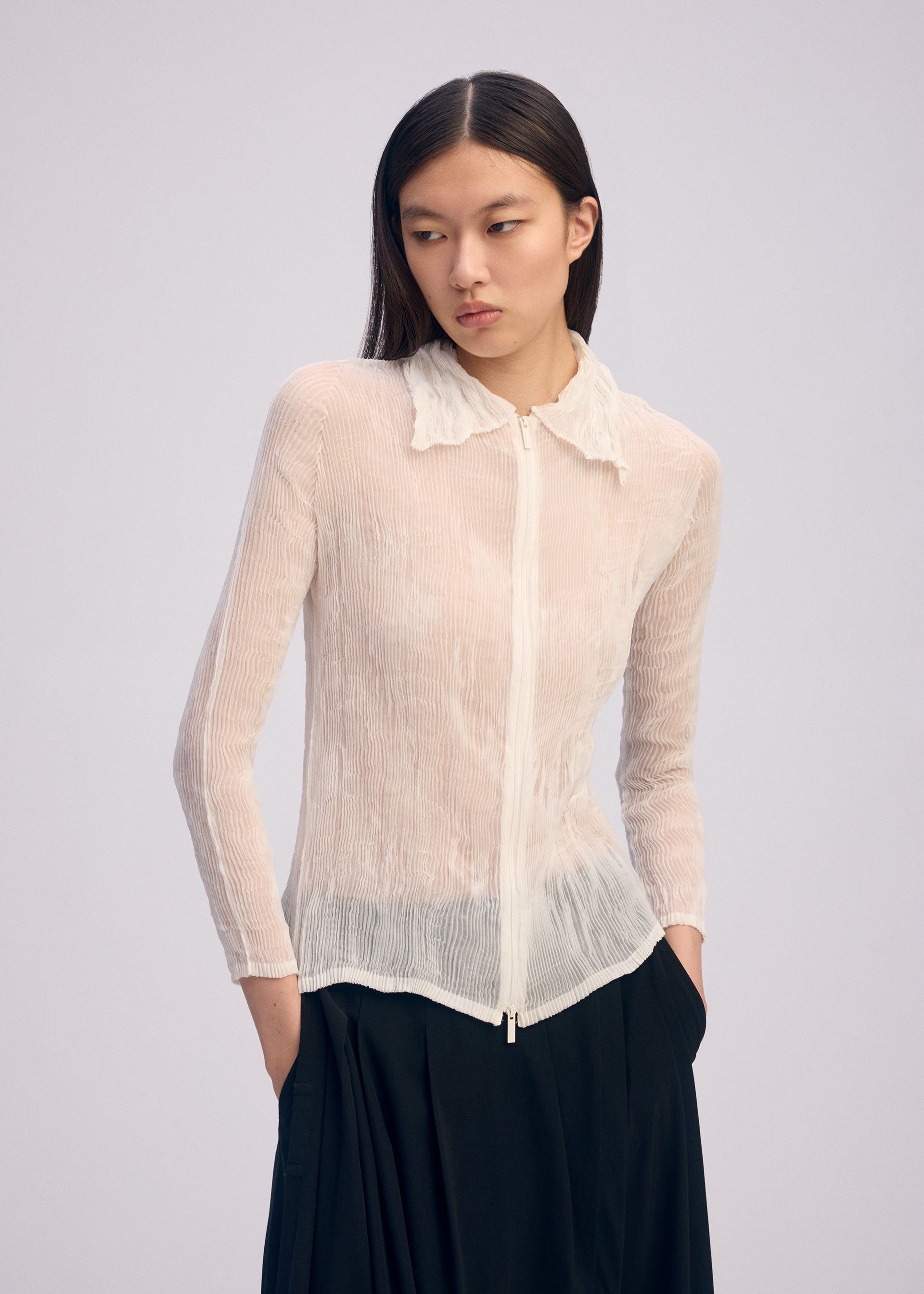 Zoomed view A person with long dark hair wears the ISSEY MIYAKE CHIFFON TWIST BK/WT-66, a sheer, textured white zip-up cardigan with a collar made from recycled polyester, and loose black pants, standing against a light background.