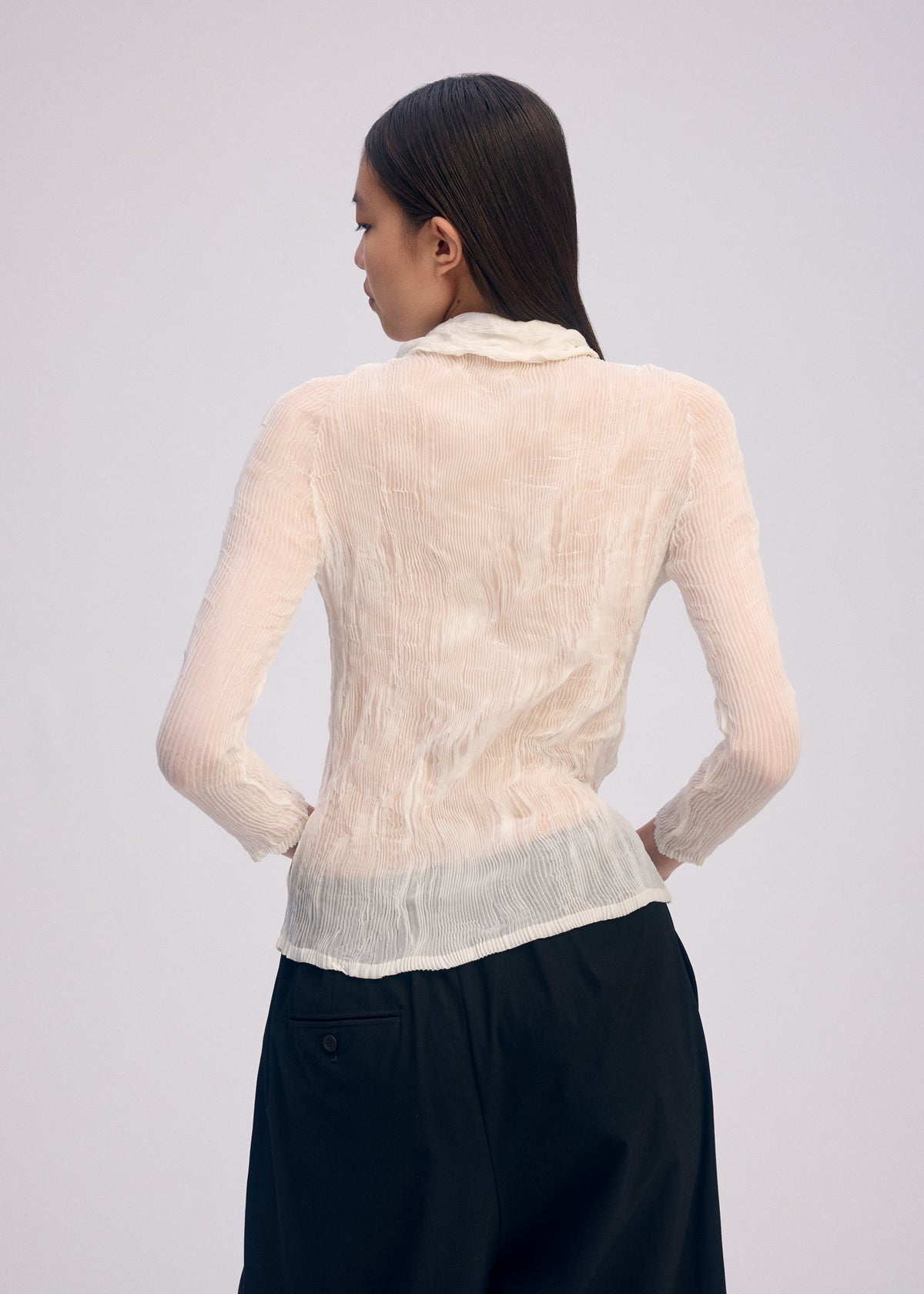A person with long, straight dark hair is seen from behind wearing the ISSEY MIYAKE CHIFFON TWIST BK/WT-66—a sheer, pleated cream top with loose black pants—against a plain light background. view 6 of 6