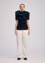 A person stands against a plain background wearing ISSEY MIYAKE's GENERIC WEAR: a structured black round-neck top with snap buttons, wide white trousers, and black shoes. They face forward with arms by their sides and a neutral expression.