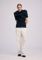 A person poses against a plain background, wearing the ISSEY MIYAKE GENERIC WEAR black round-neck top with structured shoulders, loose white trousers, and black shoes; hands behind their back and a neutral expression.