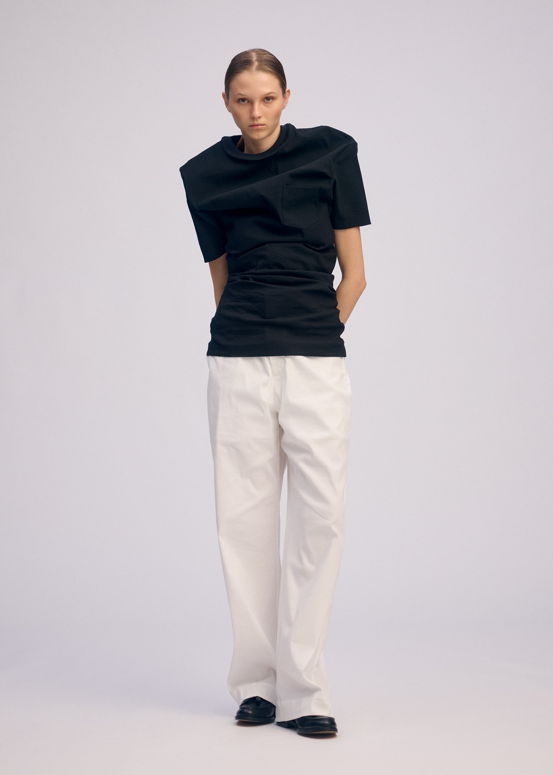 Zoomed view A person poses against a plain background, wearing the ISSEY MIYAKE GENERIC WEAR black round-neck top with structured shoulders, loose white trousers, and black shoes; hands behind their back and a neutral expression.