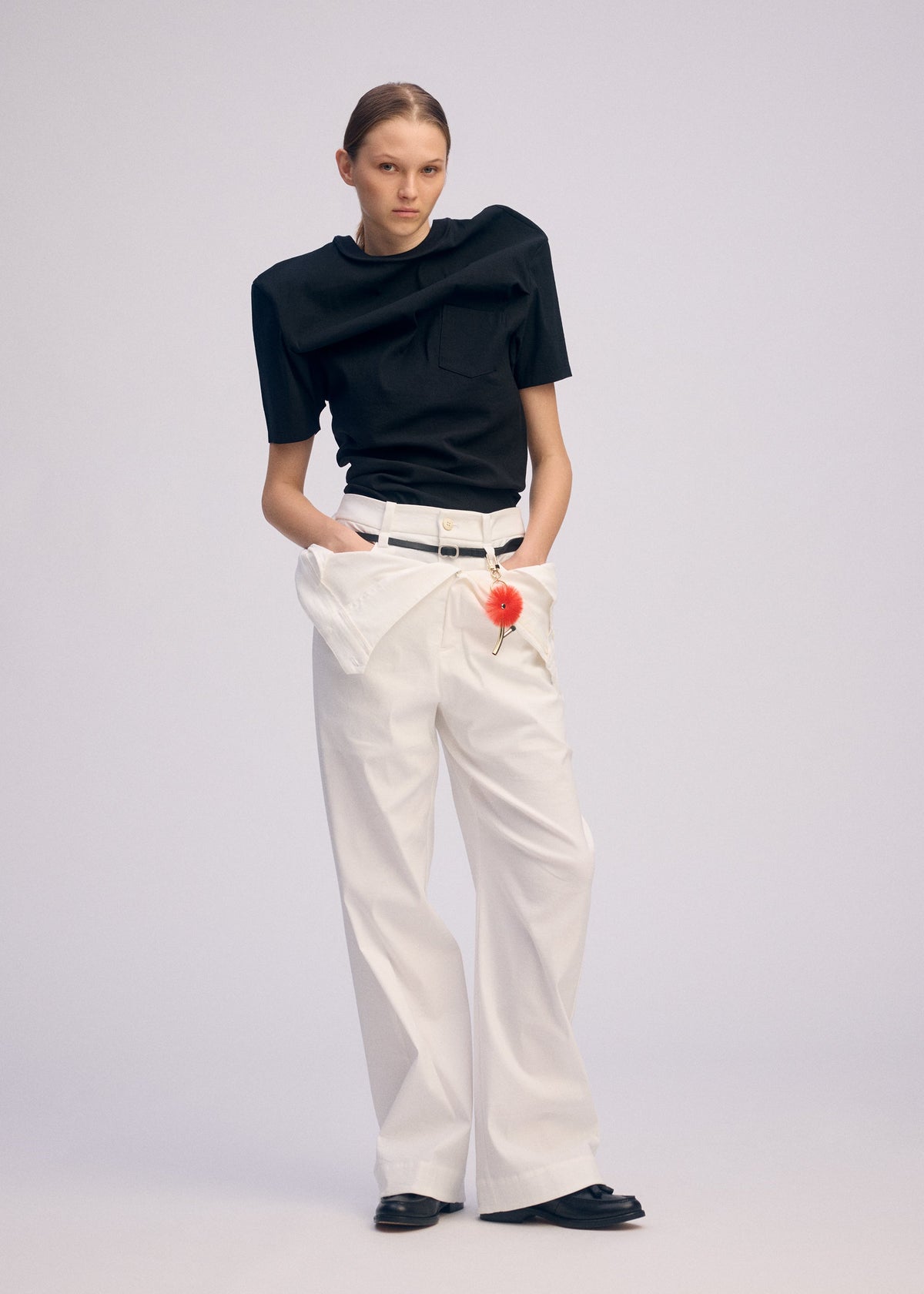 A person models ISSEY MIYAKE's GENERIC WEAR: a black round-neck top, wide-leg white trousers with pockets, black shoes, and a white belt featuring a small orange accessory, all against a plain background. view 7 of 11