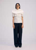 A person with straight dark hair and glasses faces forward against a light background, wearing ISSEY MIYAKE's GENERIC WEAR: a white round-neck top and loose black snap-button trousers.