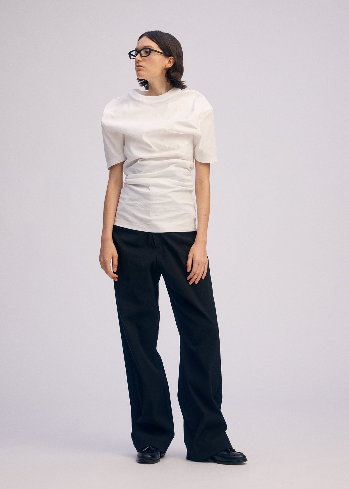 A person with shoulder-length brown hair and glasses wears the ISSEY MIYAKE GENERIC WEAR white top, loose black pants, and black shoes, standing against a plain light background. view 9 of 11