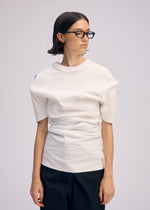 A person with short dark hair and glasses stands against a plain background, wearing ISSEY MIYAKE’s GENERIC WEAR white round-neck top with a structured design and black pants, looking to the side with a neutral expression.