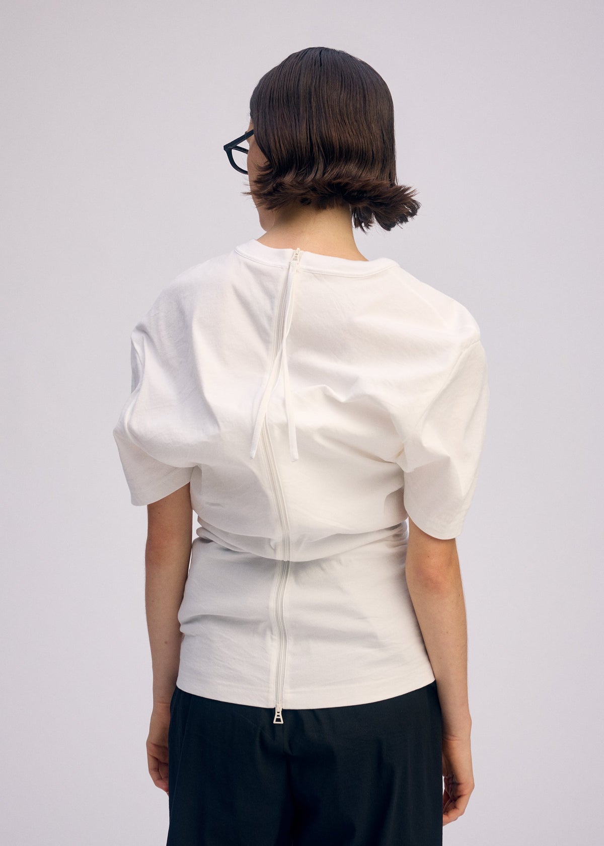 A person with short brown hair and glasses faces away, wearing ISSEY MIYAKE GENERIC WEAR—a white round-neck top with back seams, snap buttons, and drawstring—paired with dark pants against a plain light background. view 11 of 11