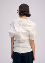A person with short brown hair and glasses faces away, wearing ISSEY MIYAKE GENERIC WEAR—a white round-neck top with back seams, snap buttons, and drawstring—paired with dark pants against a plain light background.
