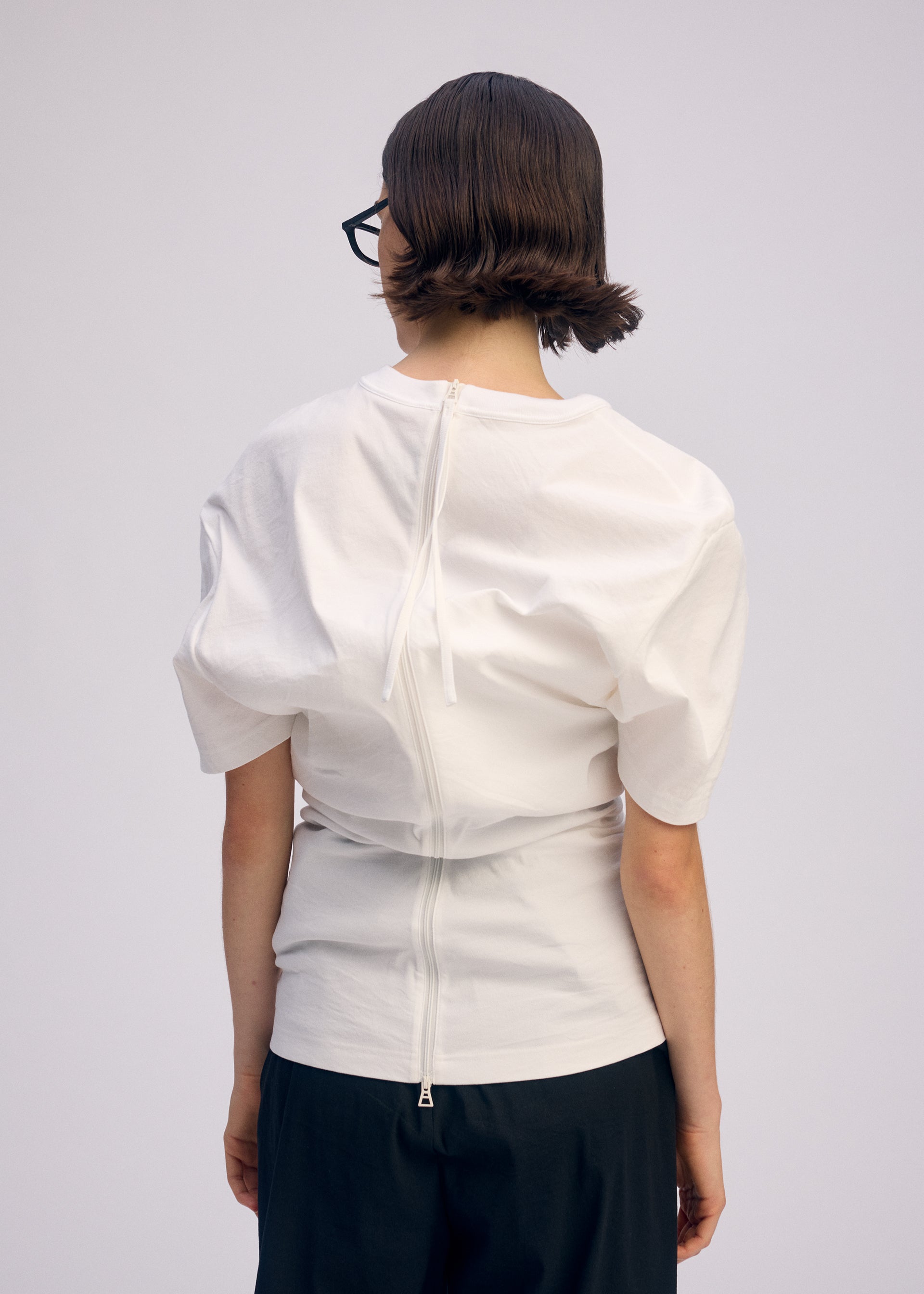 Zoomed view A person with short brown hair and glasses faces away, wearing ISSEY MIYAKE GENERIC WEAR—a white round-neck top with back seams, snap buttons, and drawstring—paired with dark pants against a plain light background.