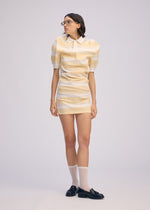 Dressed in ISSEY MIYAKE's GENERIC WEAR: STRIPES—a short, yellow-and-white striped dress with puffed sleeves—this person pairs it with glasses, sheer knee-high socks, and black loafers against a plain background.