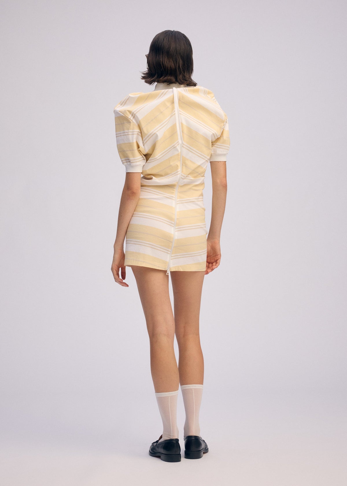 A person with short brown hair wears the ISSEY MIYAKE GENERIC WEAR: STRIPES light yellow and white striped short-sleeve dress, paired with white ankle socks and black shoes, standing against a plain light background. view 6 of 9