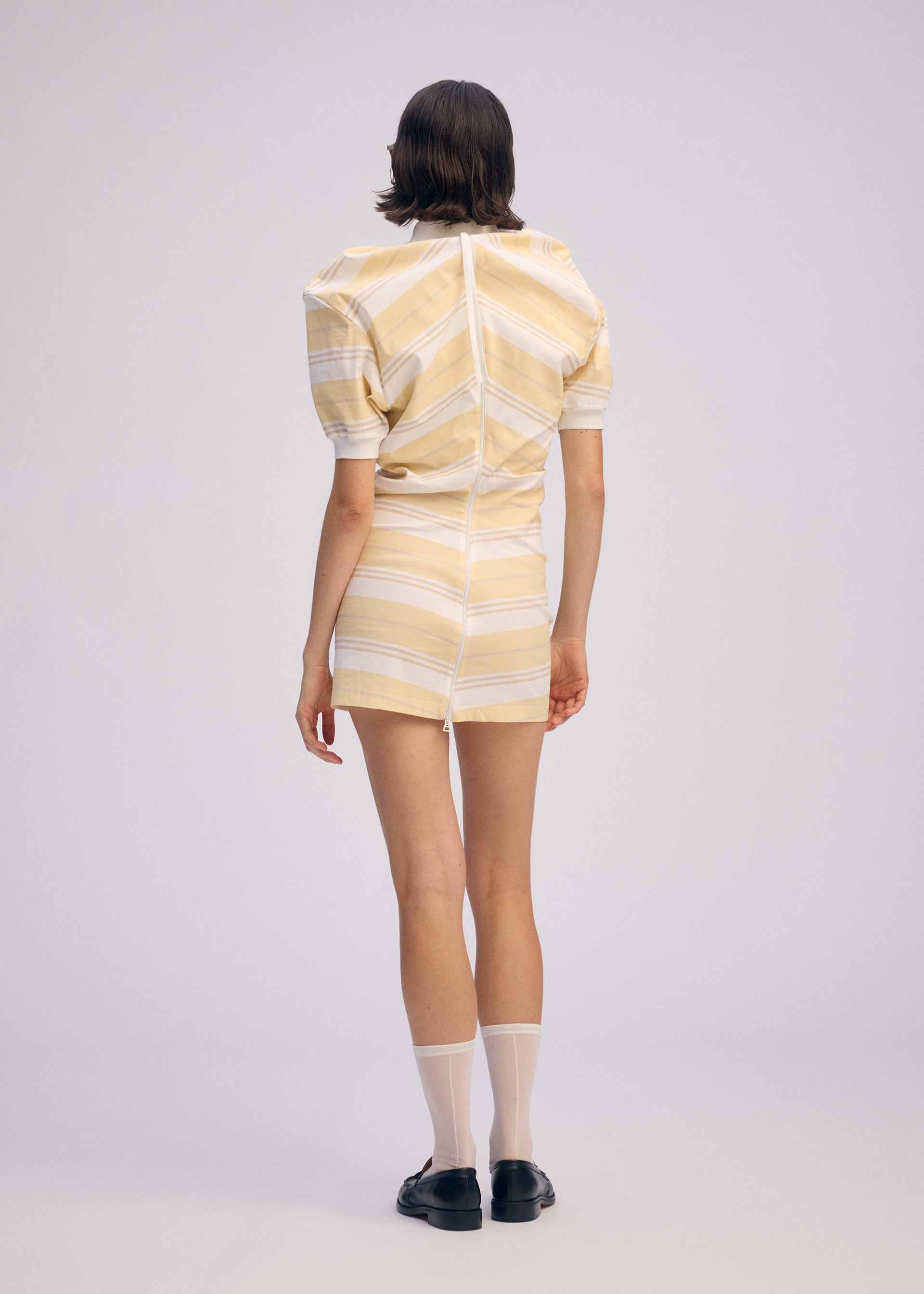 Zoomed view A person with short brown hair wears the ISSEY MIYAKE GENERIC WEAR: STRIPES light yellow and white striped short-sleeve dress, paired with white ankle socks and black shoes, standing against a plain light background.