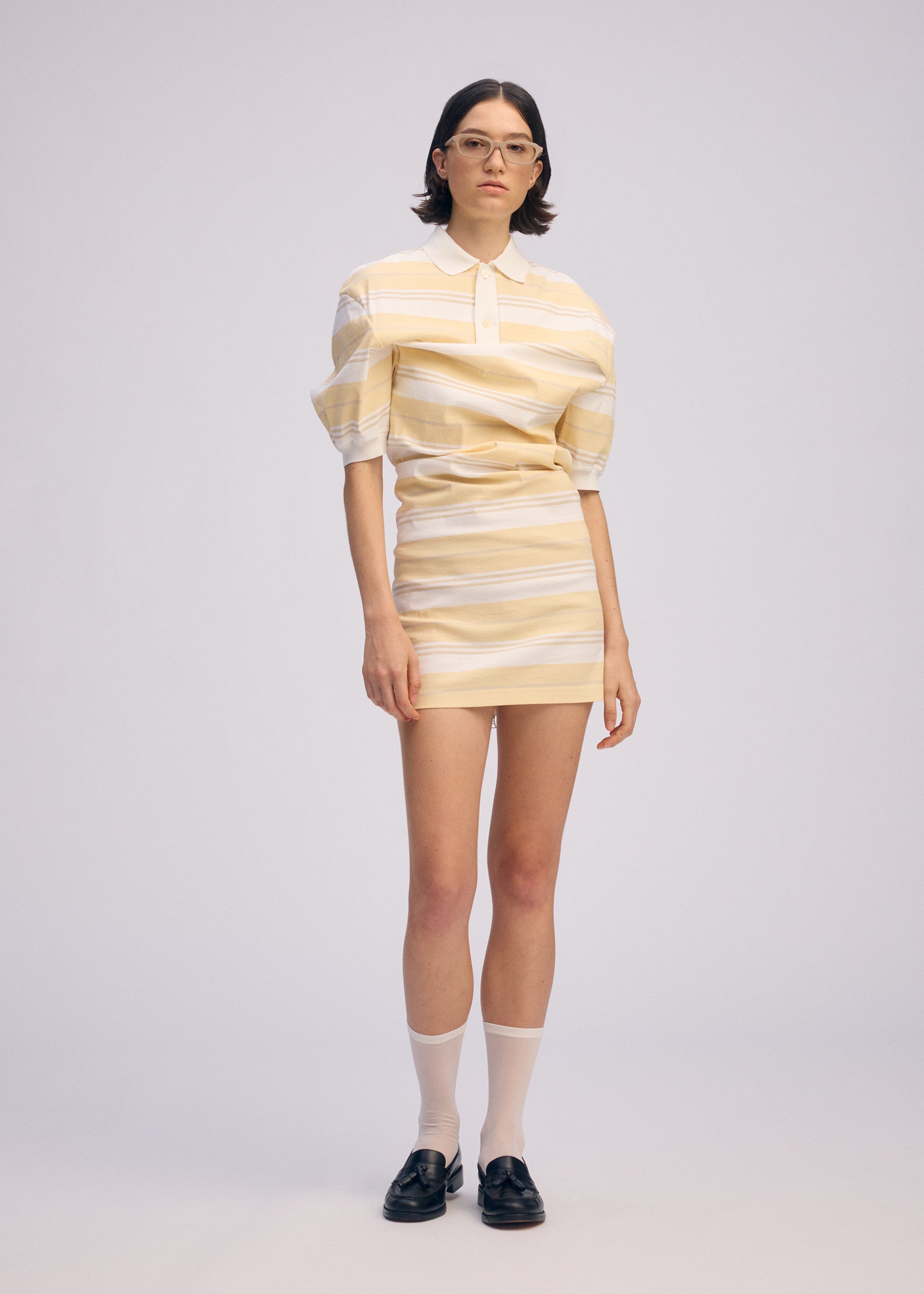 Zoomed view A person with short dark hair and glasses stands confidently against a plain light background, wearing ISSEY MIYAKE's GENERIC WEAR：STRIPES polo shirt and mini skirt, paired with white socks and black loafers.