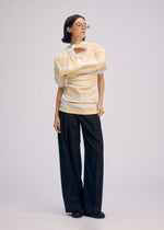 A person with short dark hair and glasses models the ISSEY MIYAKE GENERIC WEAR: STRIPES beige and white striped polo shirt, wide-leg black trousers, and black shoes against a light background, highlighting playful style.