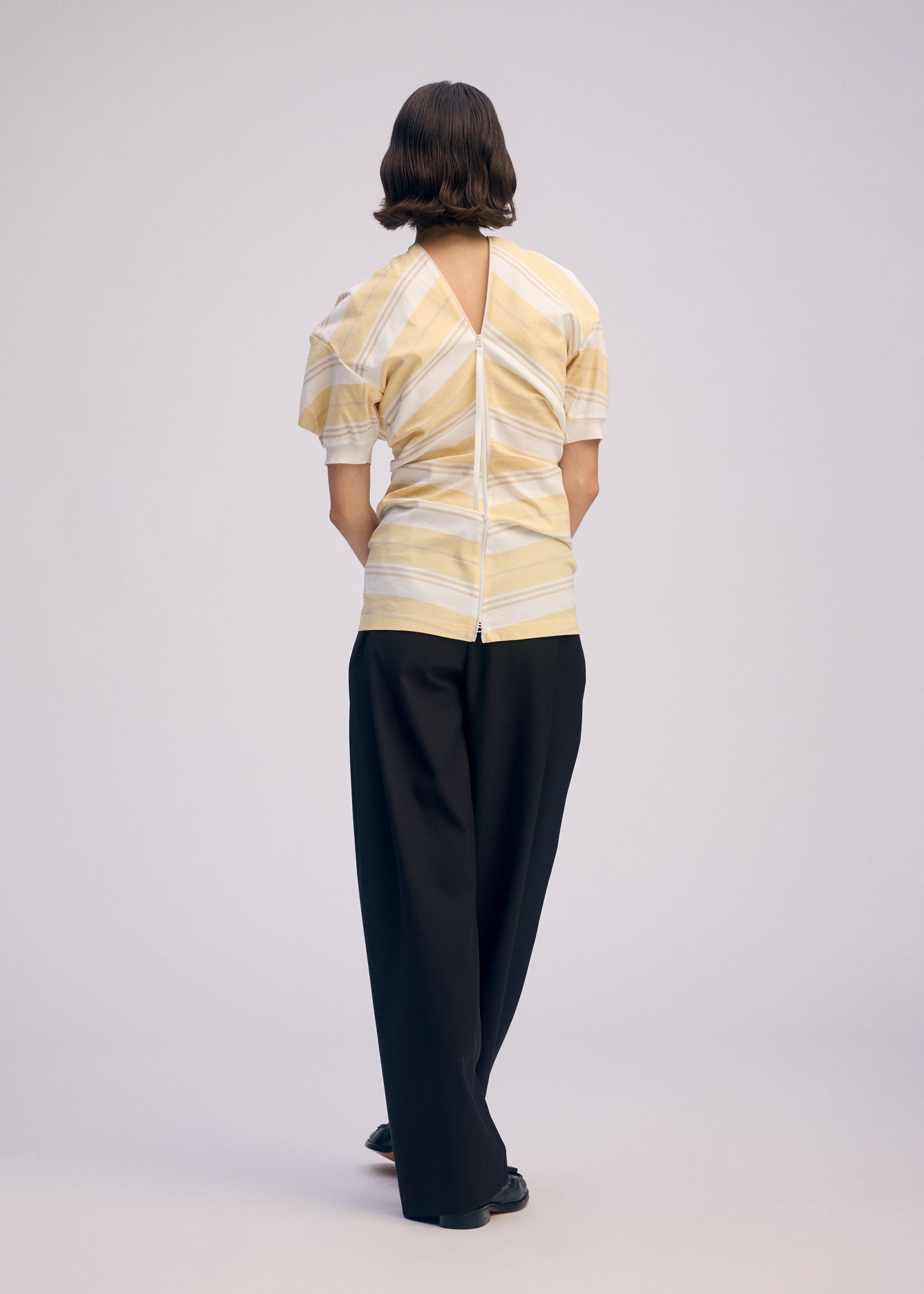 Zoomed view A person with short dark hair faces away, wearing ISSEY MIYAKE's GENERIC WEAR: STRIPES—a yellow and white striped top with a V-back cutout and zipper—paired with loose black trousers and black shoes against a plain light background.