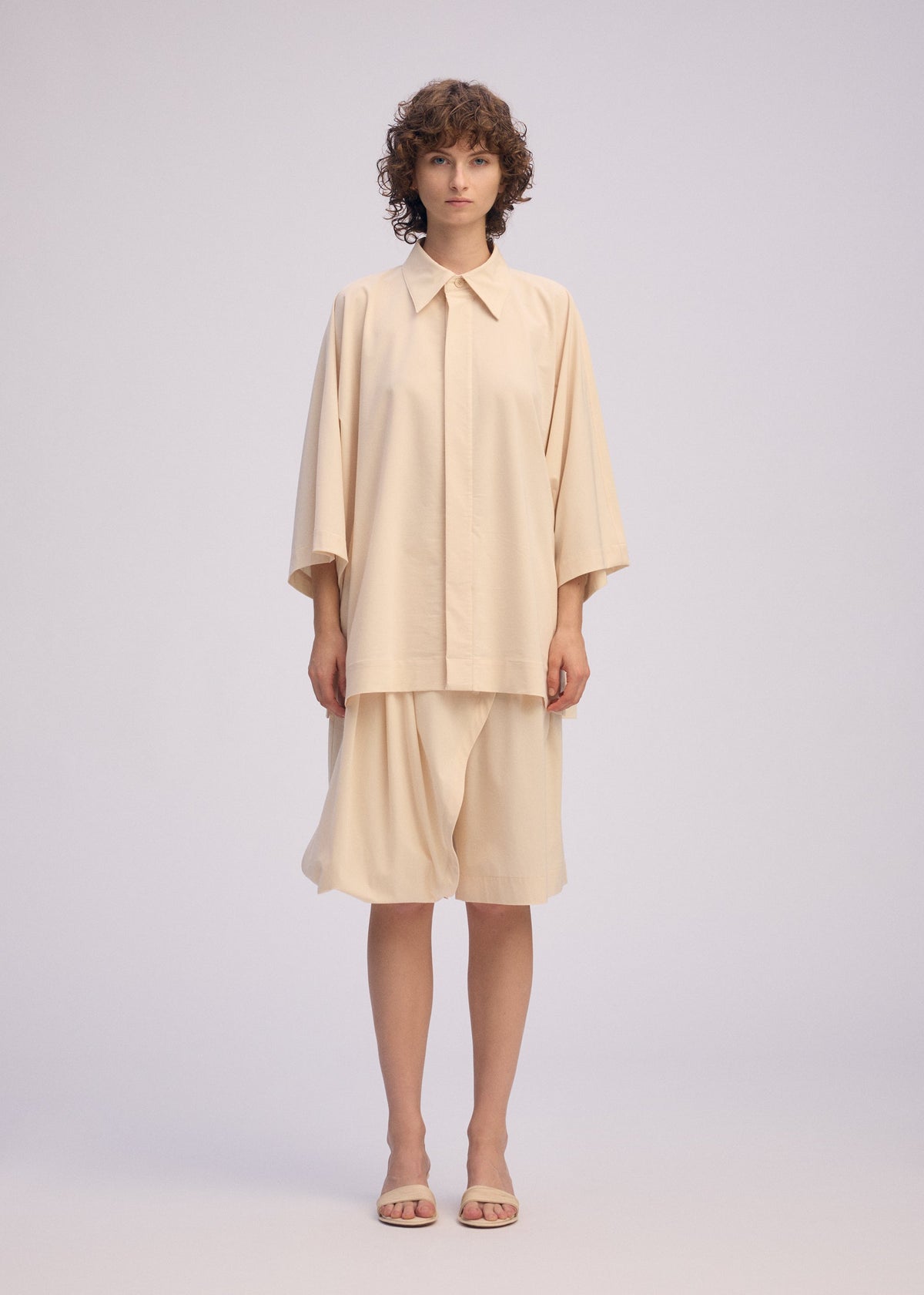 A person wearing the LOOPED shirt and matching knee-length skirt by ISSEY MIYAKE, both featuring a playful design and regular collar, stands facing forward with short curly hair and beige sandals against a plain light background. view 4 of 7