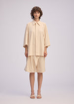 A person wearing the LOOPED shirt and matching knee-length skirt by ISSEY MIYAKE, both featuring a playful design and regular collar, stands facing forward with short curly hair and beige sandals against a plain light background.