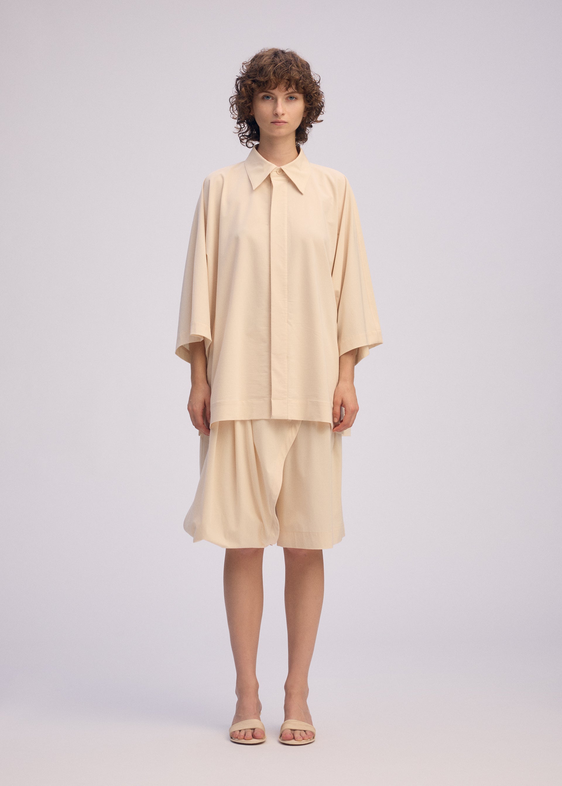 Zoomed view A person wearing the LOOPED shirt and matching knee-length skirt by ISSEY MIYAKE, both featuring a playful design and regular collar, stands facing forward with short curly hair and beige sandals against a plain light background.