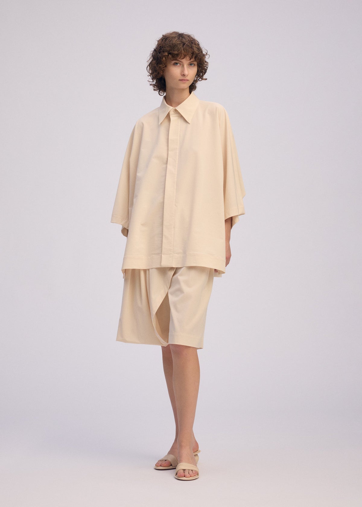 A person with short curly hair models the ISSEY MIYAKE LOOPED shirt—loose, playful design, regular collar, wide sleeves—paired with knee-length shorts and beige sandals, standing before a plain light background. view 5 of 7