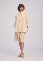 A person with short curly hair models the ISSEY MIYAKE LOOPED shirt—loose, playful design, regular collar, wide sleeves—paired with knee-length shorts and beige sandals, standing before a plain light background.
