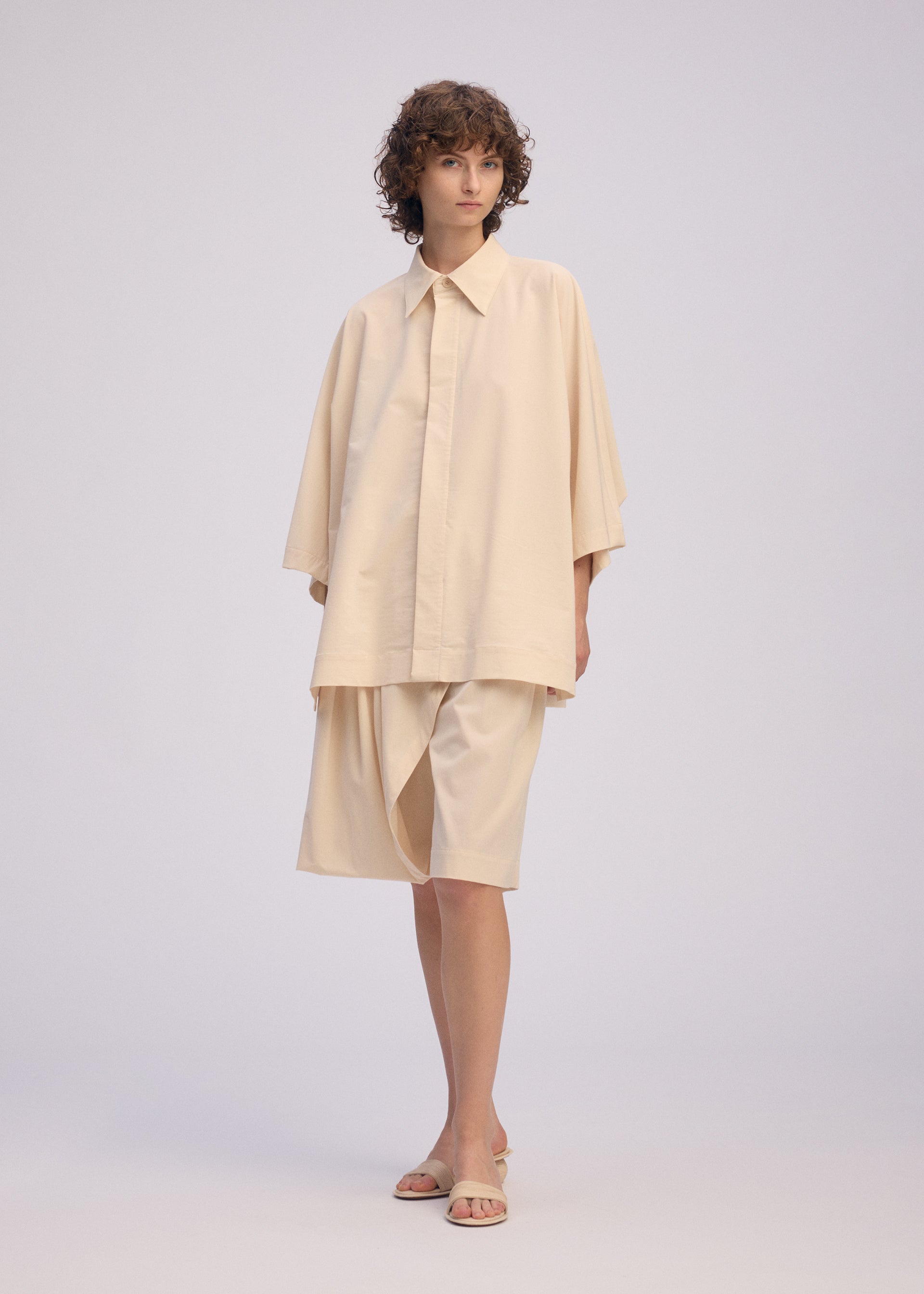 Zoomed view A person with short curly hair models the ISSEY MIYAKE LOOPED shirt—loose, playful design, regular collar, wide sleeves—paired with knee-length shorts and beige sandals, standing before a plain light background.