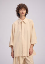 A person with short, curly brown hair wears ISSEY MIYAKE’s LOOPED set— a loose-fitting, playful shirt with wide sleeves and matching pants— standing against a plain, light background.