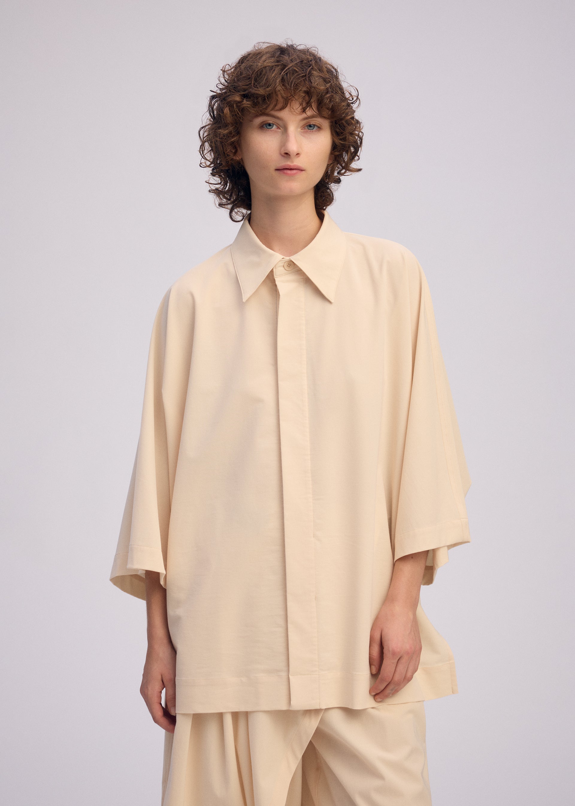 Zoomed view A person with short, curly brown hair wears ISSEY MIYAKE’s LOOPED set— a loose-fitting, playful shirt with wide sleeves and matching pants— standing against a plain, light background.