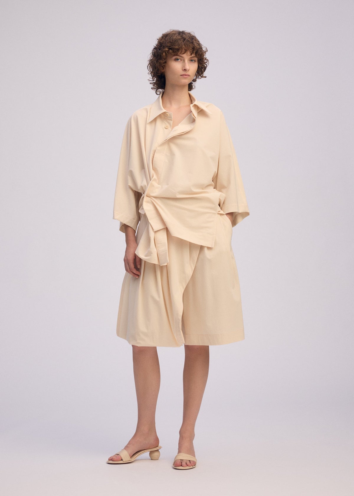 A person with short curly hair models the ISSEY MIYAKE LOOPED set, featuring a playful wide-sleeve shirt and matching knee-length skirt, paired with beige heeled sandals against a plain backdrop. view 7 of 7