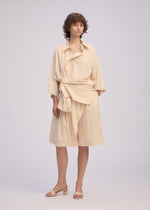 A person with short curly hair models the ISSEY MIYAKE LOOPED set, featuring a playful wide-sleeve shirt and matching knee-length skirt, paired with beige heeled sandals against a plain backdrop.