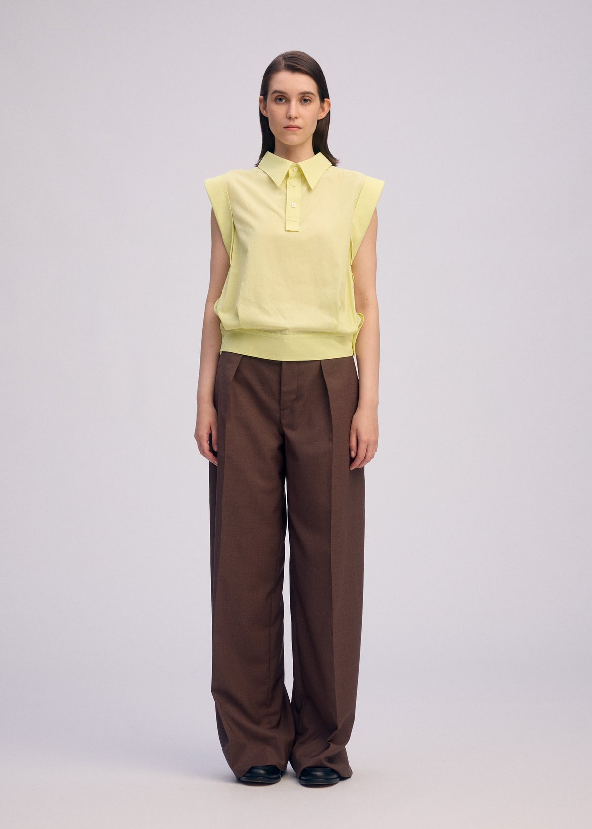 A woman faces forward in the ISSEY MIYAKE ODD SHIRT 2, a sleeveless light yellow cotton voile top, paired with high-waisted wide-leg brown trousers and black shoes, set against a plain light background. view 4 of 8
