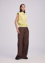 A woman with straight, shoulder-length brown hair and a neutral expression wears the ISSEY MIYAKE ODD SHIRT 2 in light yellow high-twist cotton voile, paired with wide-legged brown trousers and black shoes against a plain background—effortlessly versatile.