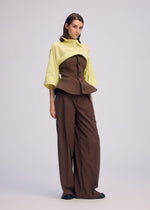 A woman with straight brown hair and a neutral expression wears wide brown trousers, a brown corset-style top, and the ISSEY MIYAKE ODD SHIRT 2— a pale yellow high-twist cotton voile shirt with an unusual draped design.