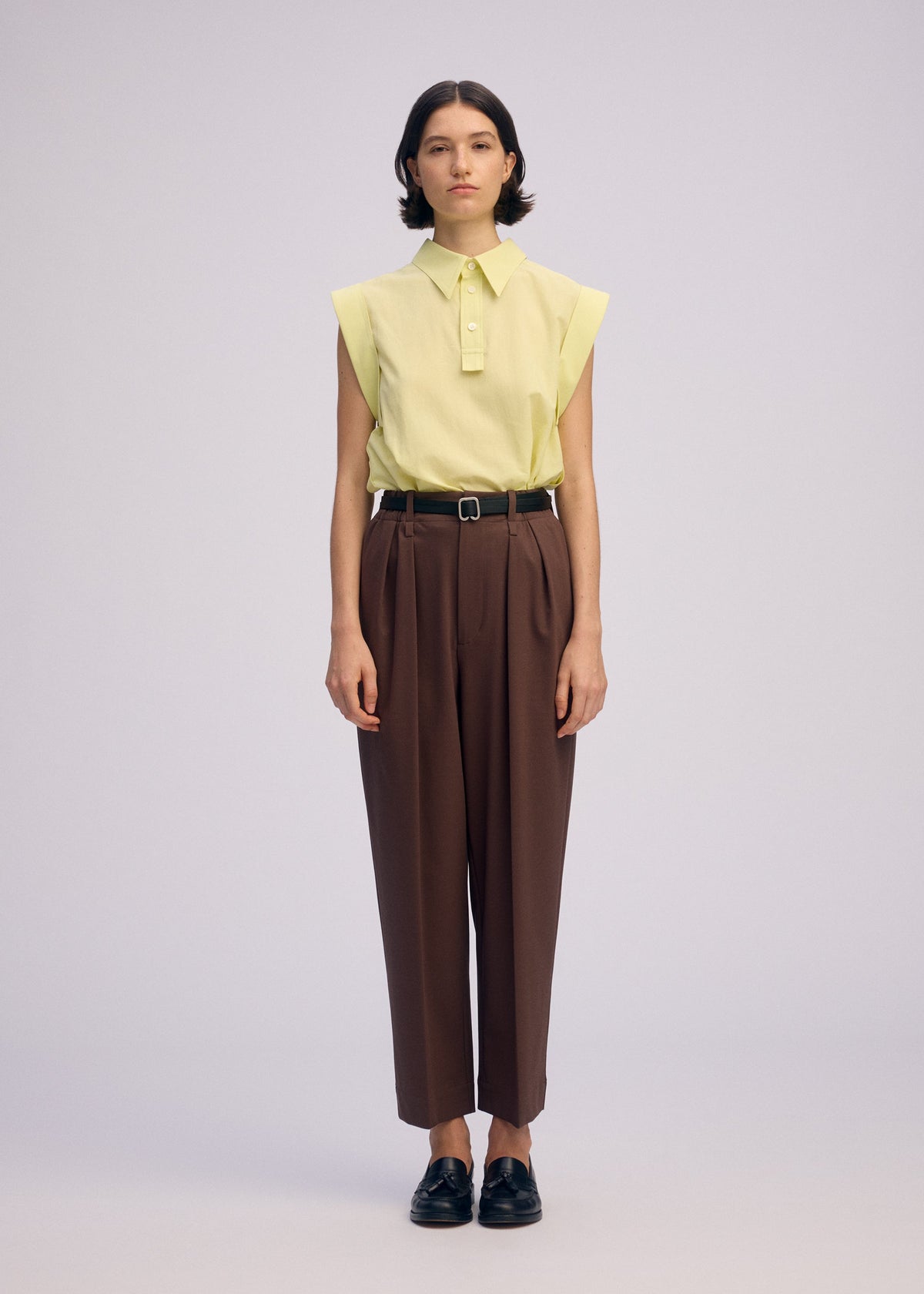A person stands facing forward in a sleeveless, light yellow ODD SHIRT 2 by ISSEY MIYAKE, tucked into high-waisted brown pleated trousers with a black belt and black loafers, set against a plain light background. view 7 of 8