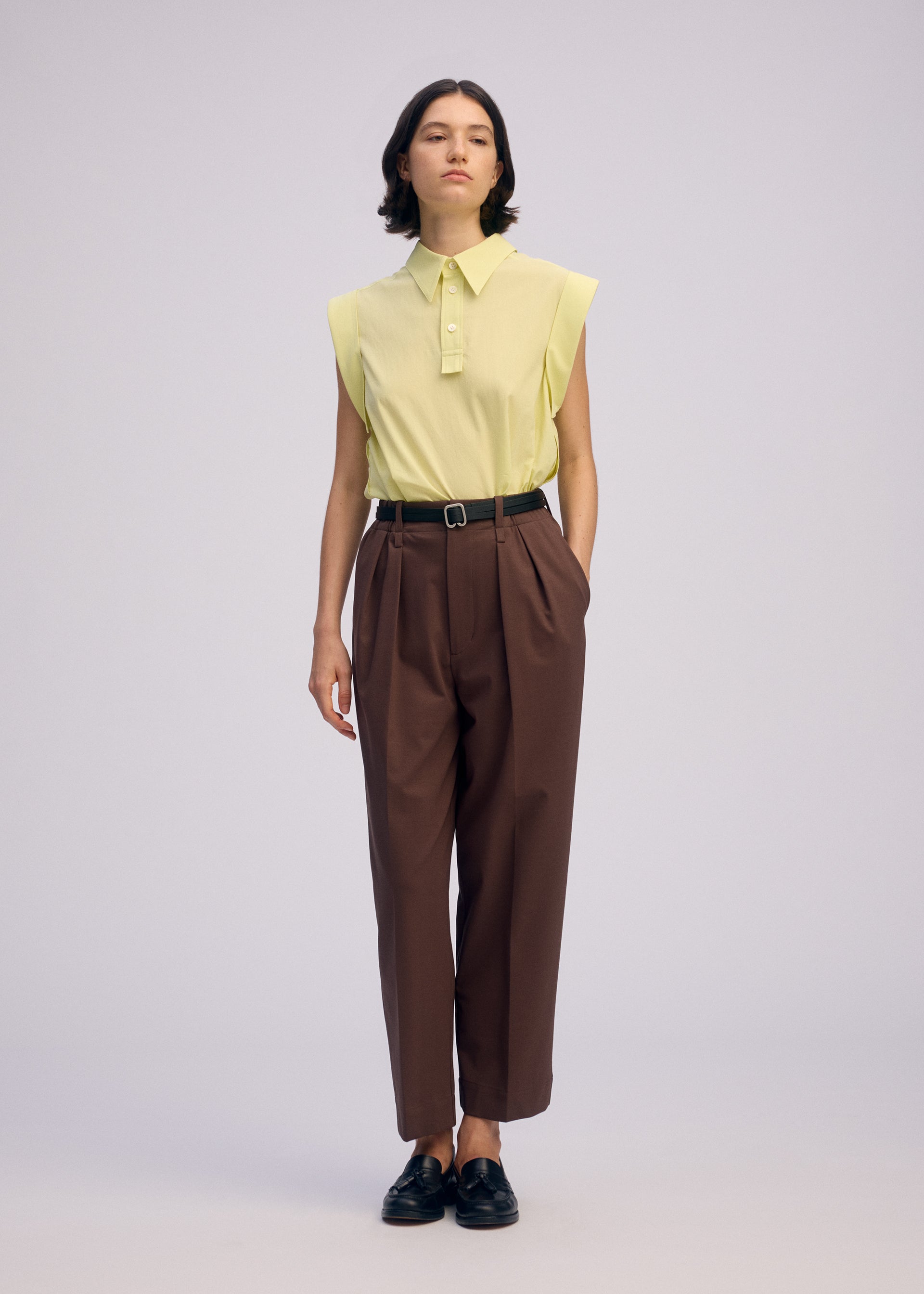 Zoomed view A person wears the ISSEY MIYAKE ODD SHIRT 2, a sleeveless yellow high-twist cotton voile shirt, tucked into high-waisted brown trousers with a black belt and loafers, hands in pockets and a neutral expression, highlighting versatile styling.
