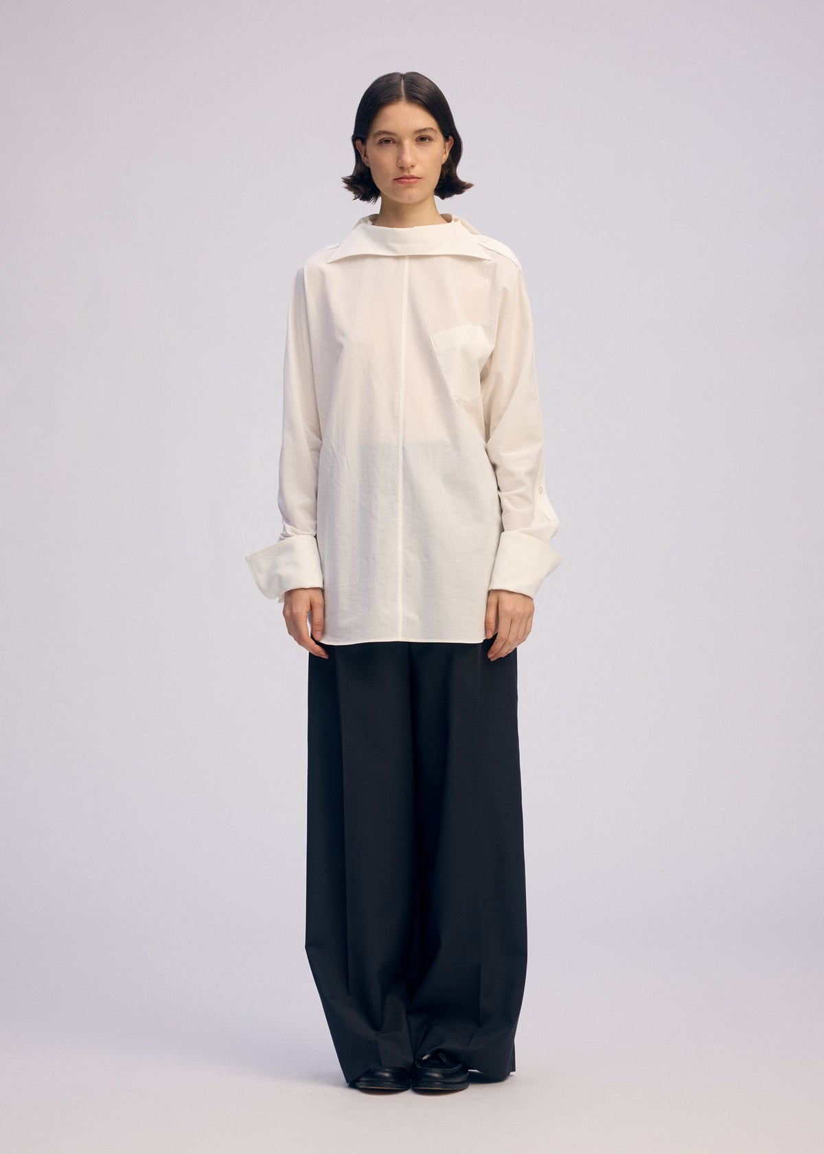 A person stands facing forward in ISSEY MIYAKE's ODD SHIRT 2, a loose white high-twist cotton voile button-up with wide sleeves, paired with oversized black trousers against a plain, light background. view 5 of 7