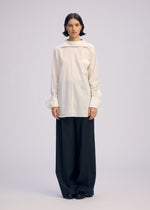 A person stands facing forward in ISSEY MIYAKE's ODD SHIRT 2, a loose white high-twist cotton voile button-up with wide sleeves, paired with oversized black trousers against a plain, light background.
