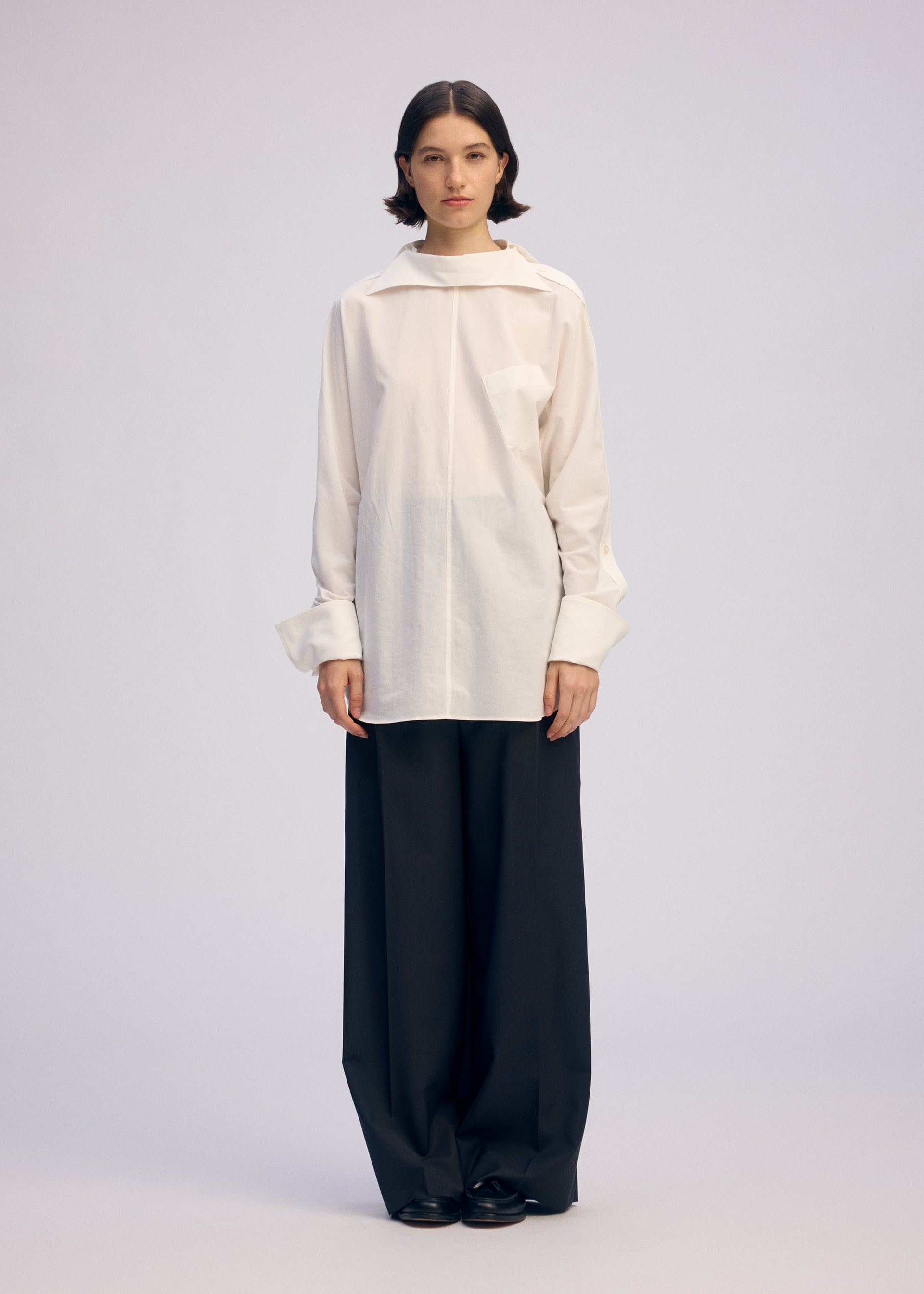 Zoomed view A person stands facing forward in ISSEY MIYAKE's ODD SHIRT 2, a loose white high-twist cotton voile button-up with wide sleeves, paired with oversized black trousers against a plain, light background.