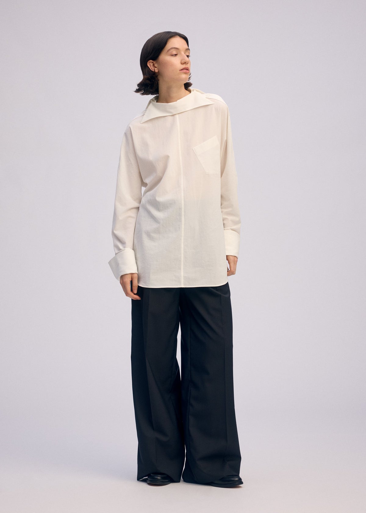 A person with short dark hair stands against a plain background, looking to the side and wearing the ISSEY MIYAKE ODD SHIRT 2—a loose white high-twist cotton voile pullover with collar and pocket—paired with wide-leg black trousers and black shoes. view 6 of 7
