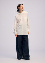 A person with short dark hair stands against a plain background, looking to the side and wearing the ISSEY MIYAKE ODD SHIRT 2—a loose white high-twist cotton voile pullover with collar and pocket—paired with wide-leg black trousers and black shoes.