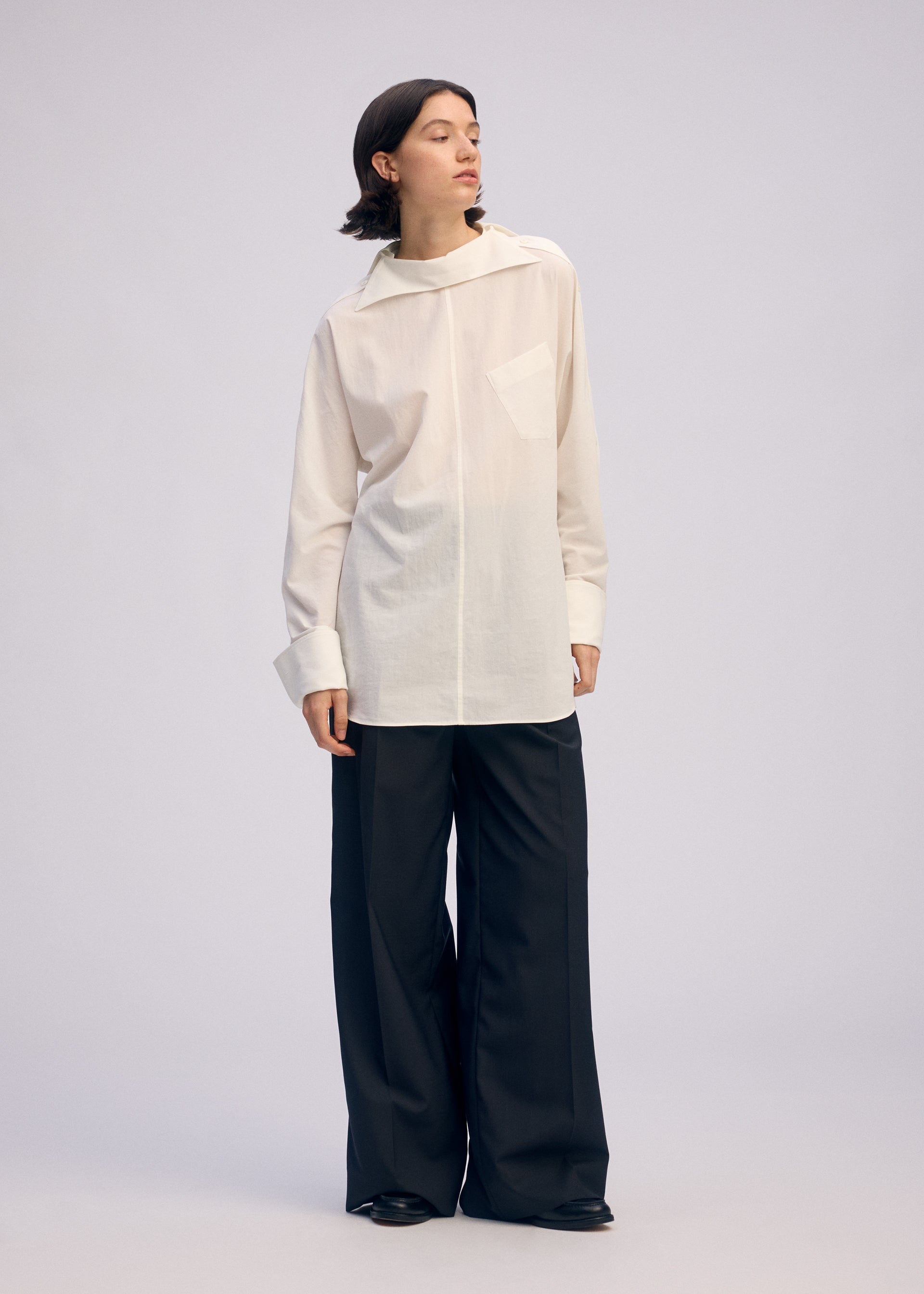 Zoomed view A person with short dark hair stands against a plain background, looking to the side and wearing the ISSEY MIYAKE ODD SHIRT 2—a loose white high-twist cotton voile pullover with collar and pocket—paired with wide-leg black trousers and black shoes.
