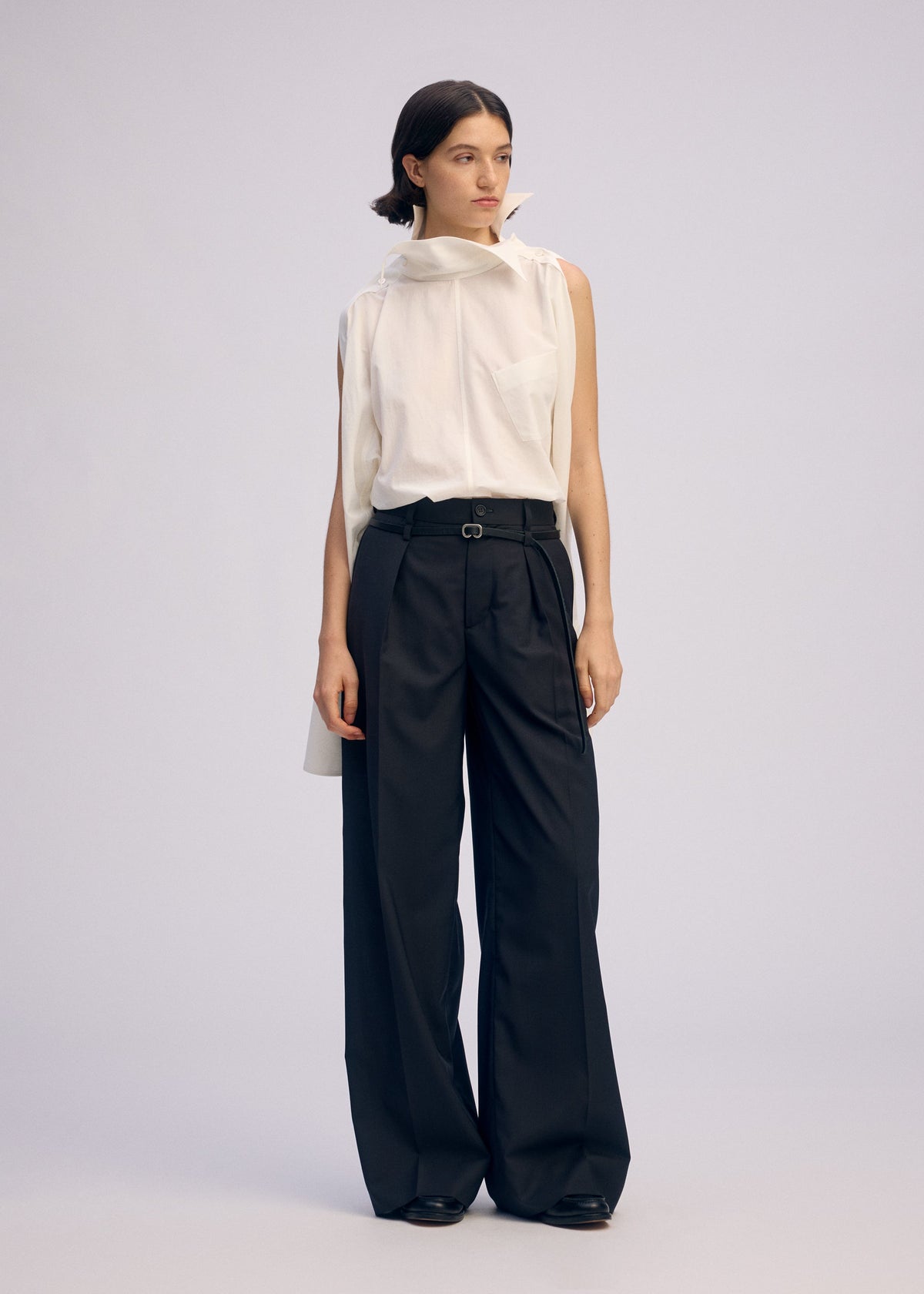 A person wears the ISSEY MIYAKE ODD SHIRT 2, a sleeveless white high-twist cotton voile pullover with an asymmetric collar, paired with black wide-leg trousers and black shoes, standing against a plain light background. view 7 of 7